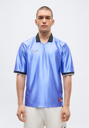 Jeune homme debout, portant un maillot de sport bleu brillant à manches courtes avec un col noir et un short blanc sur un fond uni.