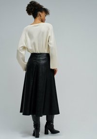 Femme aux cheveux bouclés portant un pull crème, une jupe midi noire texturée et des bottes à talons noires, se tenant devant un fond gris uni.