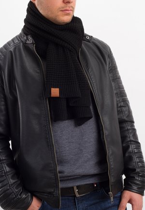 Black knitted scarf with a textured pattern and leather label, worn over a gray shirt under a black leather jacket.