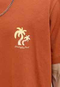Rudu t-shirt su spausdintais palmiais geltonos ir baltos spalvos, su tekstu "Huntington Beach" po piešiniu. Pagaminta iš medvilnės.