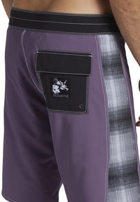 Fioletowe kąpielówki z kontrastującą czarną kieszenią z kwiatowym logo. Po bokach mają szary kraciasty wzór i białe akcenty szycia.