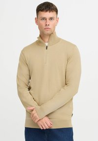 Beige halvmansjett genser laget av strikket stoff. Har lange ermer og ribbestrikkede mansjetter. Modellen står med armene krysset mot en ensfarget bakgrunn.