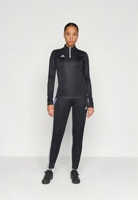 Czarny sportowy top z półzamkiem przy kołnierzu, w połączeniu z czarnymi legginsami z białymi paskami po bokach i fioletowymi akcentami. Gładka tekstura tkaniny.