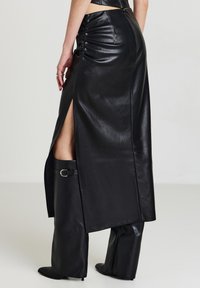 Femme portant une jupe midi en simili cuir noir avec fente latérale et des bottes à talons hautes assorties, se tenant devant un fond uni.