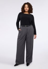 Czarna bluzka z długim rękawem z ozdobnym akcentem, zestawiona z wysokim stanem szarymi plisowanymi spodniami oraz czarnymi butami z zaostrzonym noskiem.
