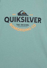Light blue cotton t-shirt featuring black and orange Quiksilver logo text, with waves and mountain graphics. Smooth texture, relaxed fit.