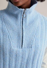 Helles Blaues, gestricktes Sweatshirt mit Reißverschluss und hohem Kragen, das über eine gerippte Textur und einen silbernen Reißverschluss auf der Vorderseite verfügt.