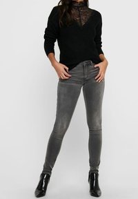 Pull noir en maille avec un col haut en dentelle, associé à un jean gris ajusté et des bottines noires, mettant en valeur une silhouette épurée.