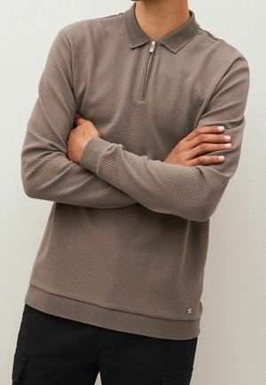 Mann mit verschränkten Armen, der ein strukturiertes braunes Langarm-Poloshirt mit Frontreißverschluss und schwarze Cargohosen trägt.