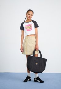 Geantă tote neagră cu un design simplu și logo, purtată cu un tricou scurt alb și negru cu un grafic colorful, asortată cu o fustă bej.
