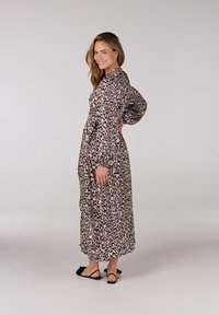 Lang, patroonjurk met een zwart en beige luipaardprint, met een relaxed fit, lange mouwen en een enkel-lange ontwerp, gecombineerd met zwarte sandalen.