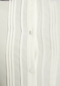 Primo piano di un tessuto bianco plissettato con pieghe verticali e due piccoli bottoni rotondi lungo una cucitura centrale.