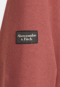 Primer plano de una tela de color óxido con una etiqueta rectangular negra de la marca Abercrombie & Fitch cosida en ella.