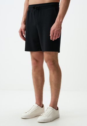Schwarze, knielange Shorts aus weichem Material, mit einem Kordelzugbund und seitlichen Taschen, kombiniert mit weißen Sneakers.