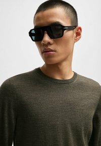 Schwarze Sonnenbrille mit rechteckiger Form und dunklen Gläsern. Getragen mit einem taillierten, strukturierten olivgrünen Pullover mit Rundhalsausschnitt.