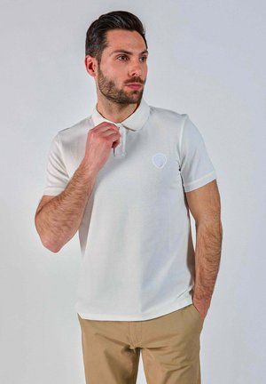 Polo blanc avec une finition texturée, doté d'un col à boutons et d'un petit logo sur la poitrine gauche ; porté avec un pantalon beige.