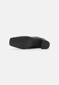 Zapato de tacón negro con suela de goma texturizada, con una punta cuadrada y material superior liso, diseñado para una estética moderna.