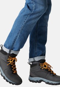 Le scarpe da trekking nere presentano lacci arancioni, una suola grigia maculata e una parte superiore testurizzata. Indossate con jeans in denim blu arrotolati.