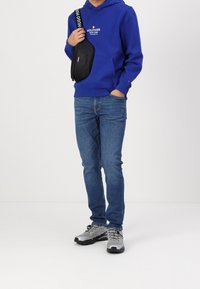 Osoba v modré mikině Tommy Hilfiger, modrých džínách, šedých teniskách a s černou Hugo crossbody taškou na bílém pozadí.