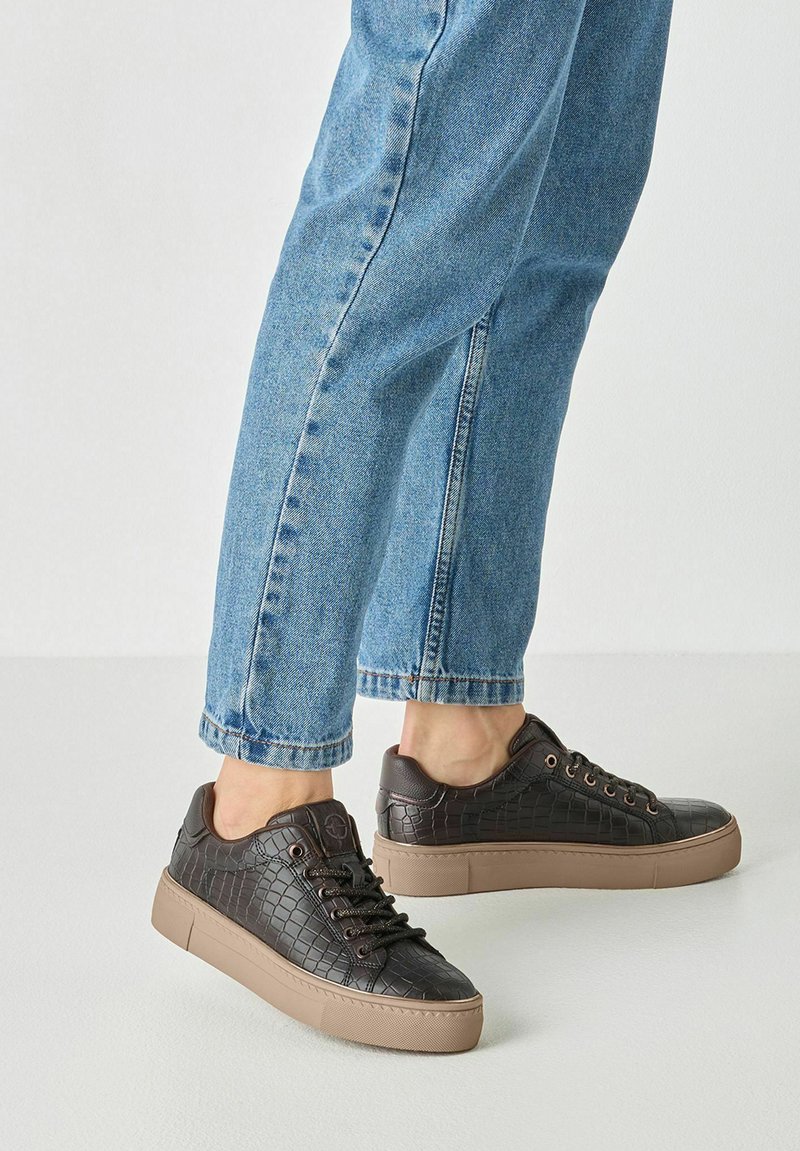 Personne portant un jean bleu et des baskets texturées marron foncé avec des semelles marron clair, debout sur un sol clair.