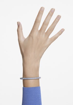 Bracelet en argent au design torsadé, ornée de petites pierres précieuses transparentes. Porté sur un poignet fin, associé à un pull bleu côtelé.
