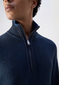Marineblauer Strickpullover mit hohem Kragen und halbem Reißverschluss, ausgestattet mit einem strukturierten Rippenmuster und kontrastierender Innenseite.