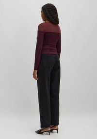 Langärmliges, burgunderfarbenes Oberteil mit durchscheinenden Schulterdetails, kombiniert mit hochgeschnittenen, schwarzen, weiten Jeans und schwarzen, spitzen Pumps.