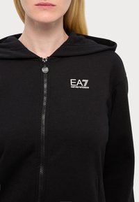 Felpa nera con zip, realizzata in un tessuto morbido, con chiusura frontale a zip e logo EA7 ricamato in bianco nella parte sinistra del petto.