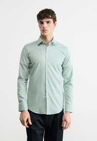 Νέος άνδρας που φοράει πουκάμισο μακρυμάνικο, ανοιχτού πράσινου χρώματος με κουμπιά και μαύρο παντελόνι, όρθιος μπροστά σε απλό λευκό φόντο.