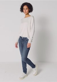 Vaaleanharmaa neulepusero, jossa on leveä kaula-aukko, yhdistetty sinisiin kapealinjaisiin farkkuihin ja valkoisiin tennareihin. Sileä pinta, rento istuvuus, minimaalinen yksityiskohtaisuus.