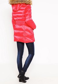 Veste matelassée rouge vif avec une capuche en fourrure, présentant un design matelassé et des poches latérales, assortie à un jean skinny bleu foncé et des bottines.