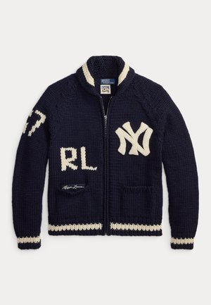 Jeune homme debout les mains dans les poches, portant un pull zippé bleu marine avec des lettres blanches, une chemise en denim clair, un haut rayé, un pantalon blanc et des baskets.