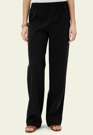 Frau trägt schwarze Hose mit weitem Bein und elastischem Bund sowie braune Sandalen mit offenen Zehen, steht vor hellem Hintergrund.