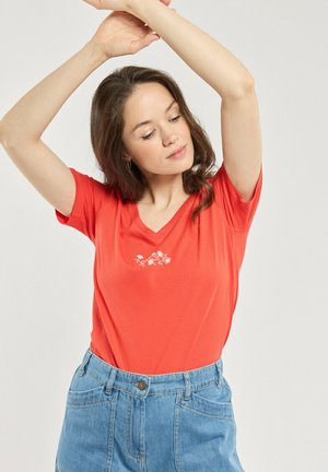 Femme aux longs cheveux bruns portant un t-shirt rouge à col en V avec un petit motif floral blanc et un jean bleu taille haute, levant les bras au-dessus de la tête.