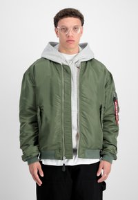 Veste bomber verte avec une finition brillante, des poignets côtelés et une poche zippée avec une étiquette rouge. Portez au-dessus d'un sweat à capuche gris.
