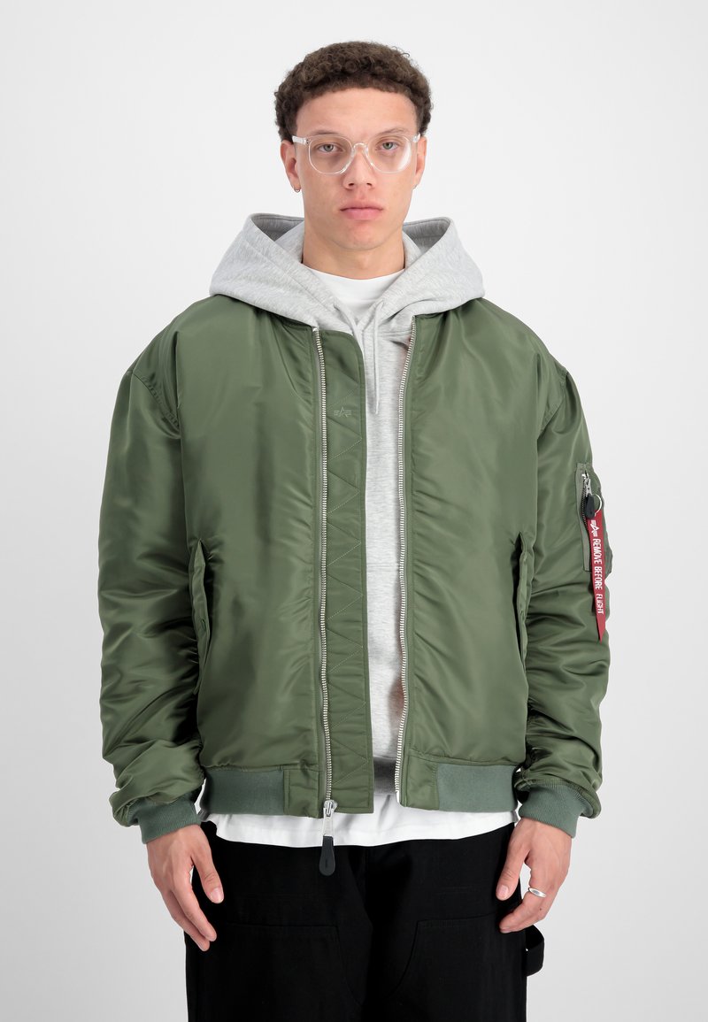 Veste bomber verte avec une finition brillante, des poignets côtelés et une poche zippée avec une étiquette rouge. Portez au-dessus d'un sweat à capuche gris.