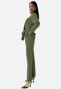 Olivgrünes zweiteiliges Outfit mit einem langärmligen Oberteil, das an der Taille gebunden wird, und passenden, eng anliegenden Hosen, ergänzt durch schwarze High Heels.