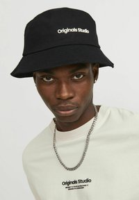 Black bucket hat with the text "Originals Studio" in white, featuring a wide brim and textured fabric. Worn with a light shirt and chain necklace.