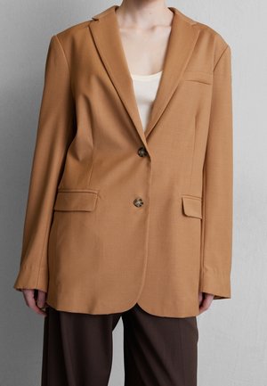 Personne portant un blazer couleur chameau à deux boutons avec des poches à rabat, par-dessus un haut blanc et un pantalon marron foncé, debout contre un mur gris.