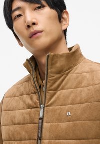 Brūns zamšs veste ar augstu apkakli, kas satur quiltu daļas, sudraba rāvējslēdzēju un mazu metāla logotipu uz krūtīm.