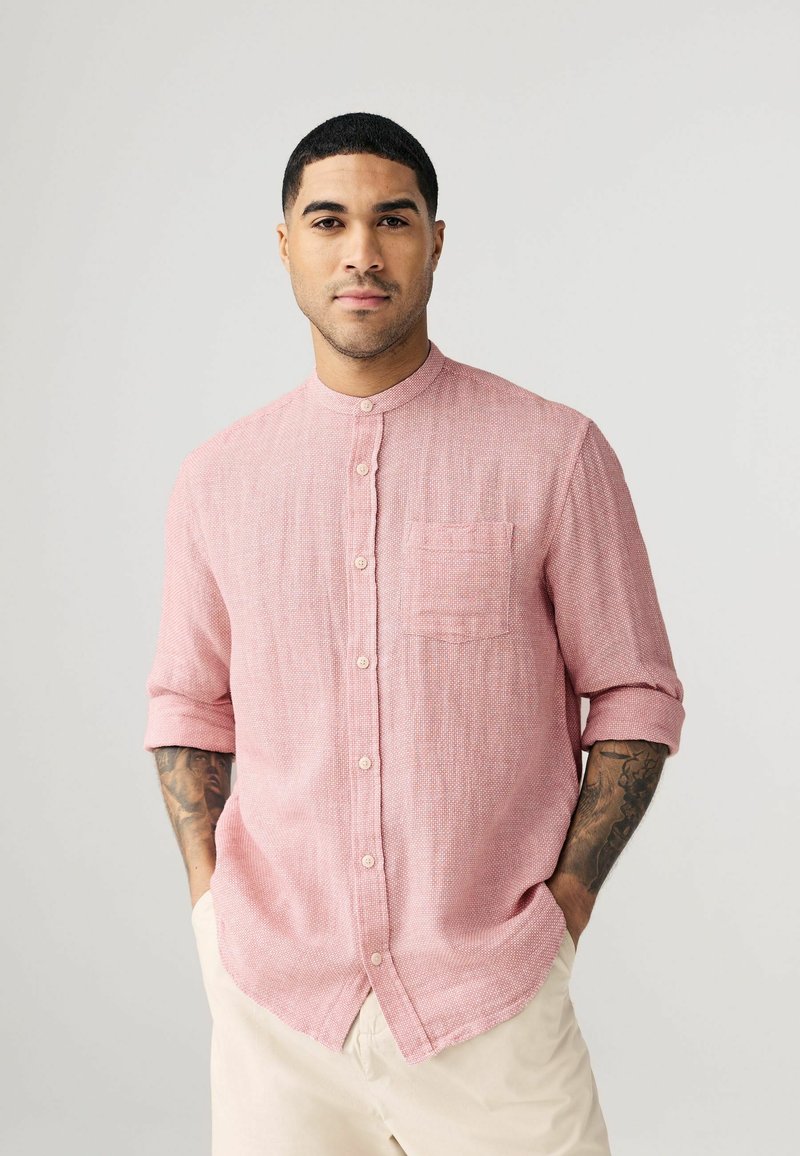 Camisa de botones de color rosa claro con cuello mandarín, mangas cortas enrolladas y un bolsillo en el pecho, que presenta un patrón de tejido texturizado.