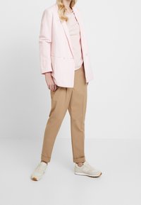 Heller rosa Leinenblazer mit zwei Knöpfen, kombiniert mit einem beigen Top und beigen, schmal zulaufenden Hosen. Die Schuhe sind beigen Sneakers mit weißen Akzenten.