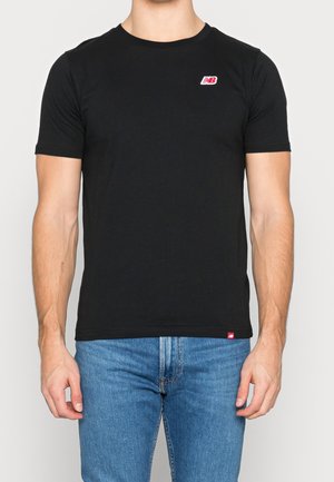 Mand iført en simpel sort kortærmet t-shirt med små røde og hvide New Balance-logoer og blå jeans, stående mod en hvid baggrund.