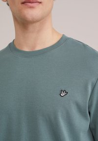 Man draagt een teal T-shirt met ronde hals en een klein zwart-wit vogellogo op de linkerborst, tegen een effen lichte achtergrond.