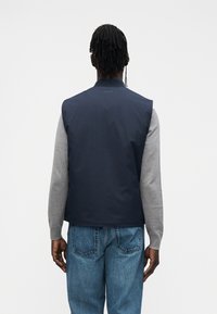 Sötétkék mellény szürke ujjakkal, sima anyagból készült. Magas gallérral és egyszerű dizájnnal rendelkezik. A hátoldalról minimális márkajelzés látható.