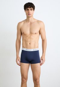 Νέος άνδρας που στέκεται όρθιος φορώντας μπλε σκούρα μπόξερ με λευκή ζώνη με το λογότυπο GANT σε μονόχρωμο φόντο.