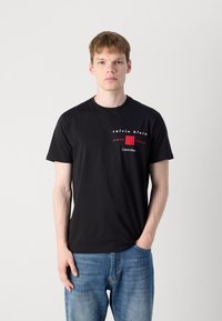 Zwart katoenen T-shirt met ronde hals, korte mouwen en een logo op de voorkant in witte en rode tekst. Gekleed met blauwe spijkerbroek.