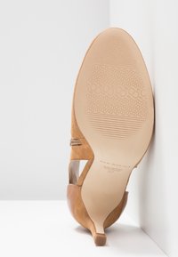 Chaussure à talon haut en daim beige inclinée vers le haut, montrant la semelle texturée avec le nom de la marque, la taille 37, et un design à bout pointu.