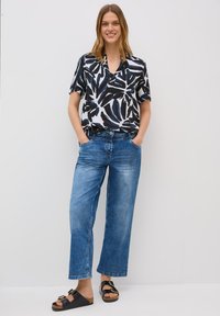 Lächelnde Frau mit langen Haaren steht vor einem weißen Hintergrund und trägt ein schwarz-weiß gemustertes Hemd, legere blaue Jeans und schwarze Sandalen.