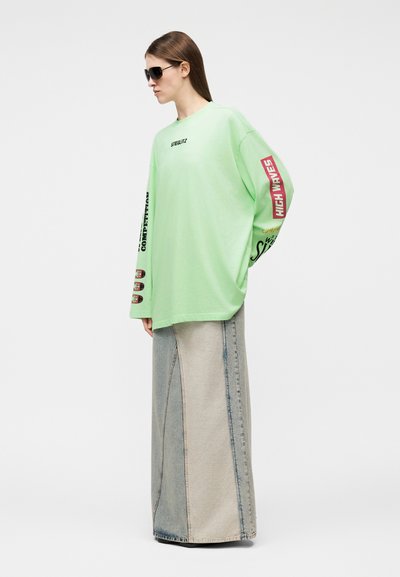 Γυναίκα με μαύρα γυαλιά ηλίου, φαρδιά ανοιχτοπράσινη μπλούζα με μακριά μανίκια και κείμενο, και μακριά φούστα από τζιν σε πατς, στέκεται προφίλ.